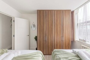 A bedroom with two beds, a wardrobe, a window with sheer curtains, and a wall-mounted clock. - Bright 1-Bedroom Apartment with Free Parking (London)