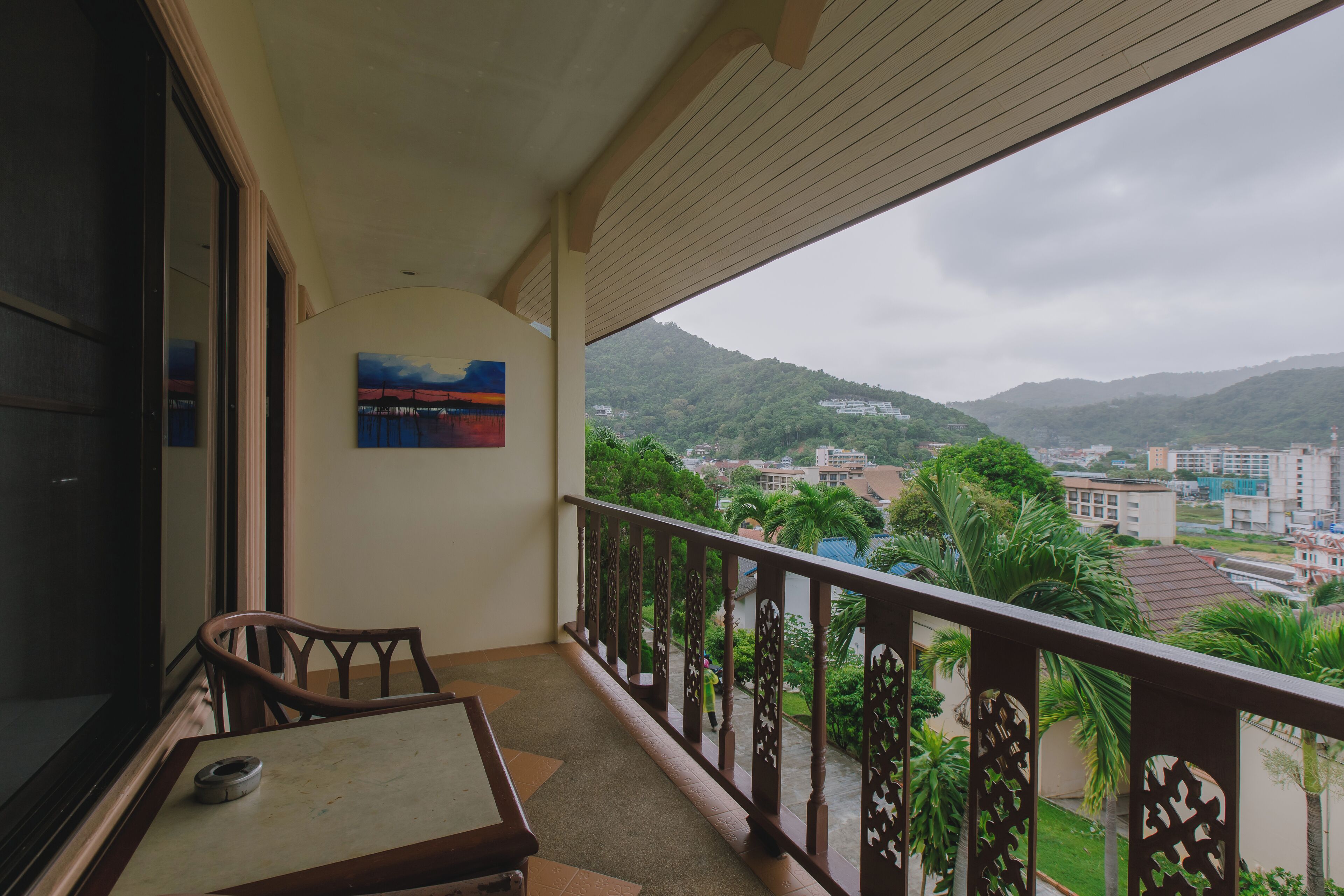A balcony with a table and chairs, a painting on the wall, and a view of a cityscape and mountains.