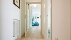 Un couloir avec un radiateur, une porte donnant sur une pièce où se trouve un canapé bleu, et un tableau encadré accroché au mur.