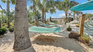 Un hamac est installé sur une plage de sable, avec des palmiers et une clôture en bois en arrière-plan.