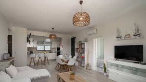 Un salon moderne avec un canapé, une table basse, un téléviseur et un coin repas comprenant des chaises, et une cuisine en arrière-plan.