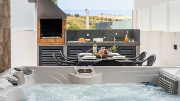 Un bain à remous doté d’un panneau de commande, une table avec des chaises, un barbecue et une vue sur le paysage.