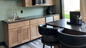 A modern kitchen with wooden cabinets, a stainless steel sink, and a black countertop. There are two black bar stools and a window with curtains.