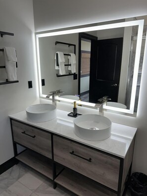 A modern bathroom with two round sinks, a large mirror with integrated lighting, and a dark door in the background.