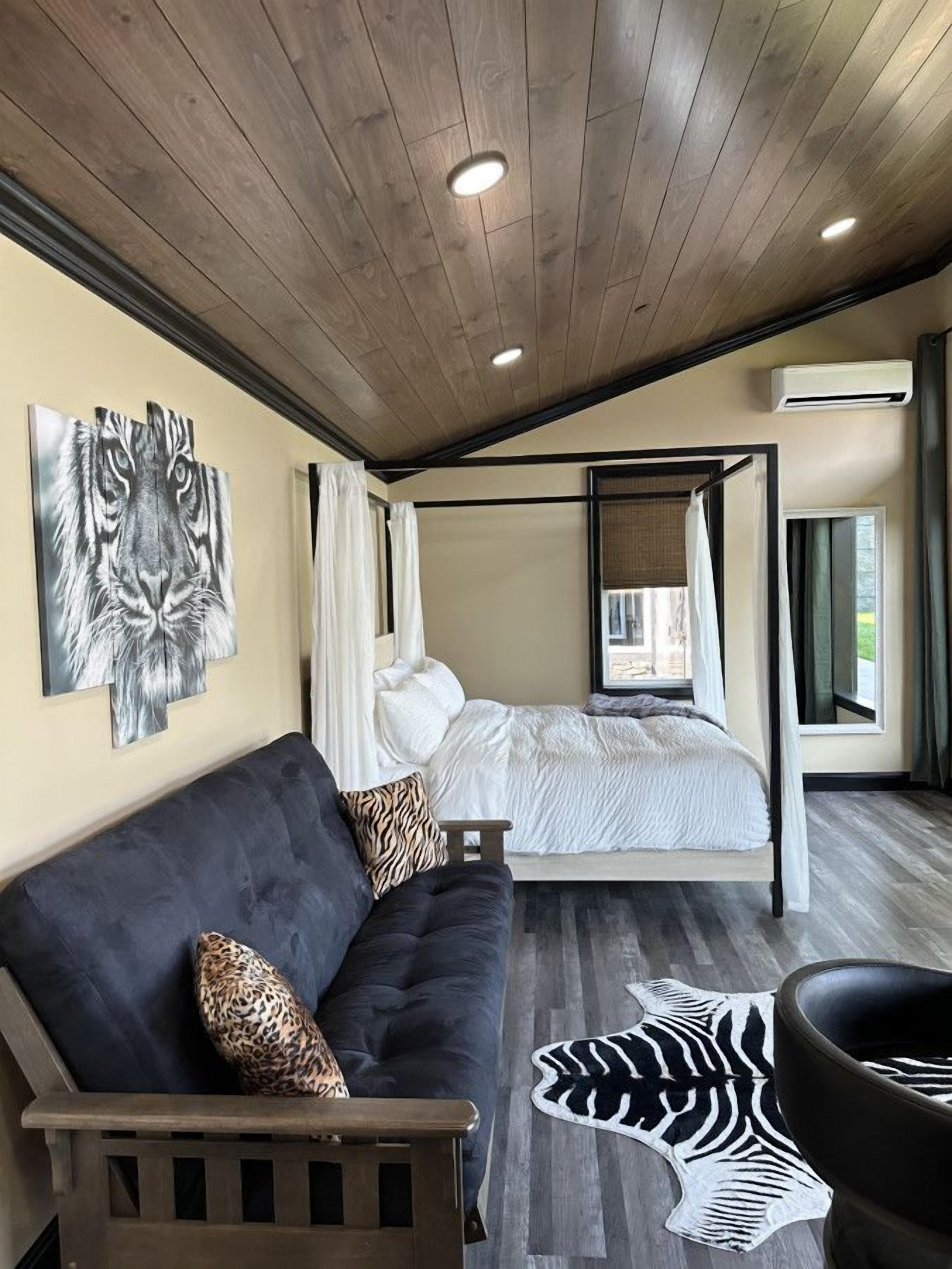 Et moderne soveværelse med træloft, seng med hvide sengetøj, mørkeblå sofa med leopardprint puder og en måtte med zebra-mønster.