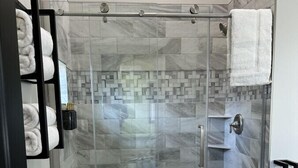 A glass-enclosed shower with a tiled floor and walls, a towel rack with towels, and a toilet paper holder on the wall. - Big Cat Bungalows at The Haven - Tiger 4 (Chittenango)