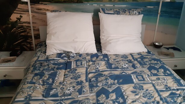 Uma cama com colcha estampada de flores, almofadas brancas e vista para a praia através de uma janela.