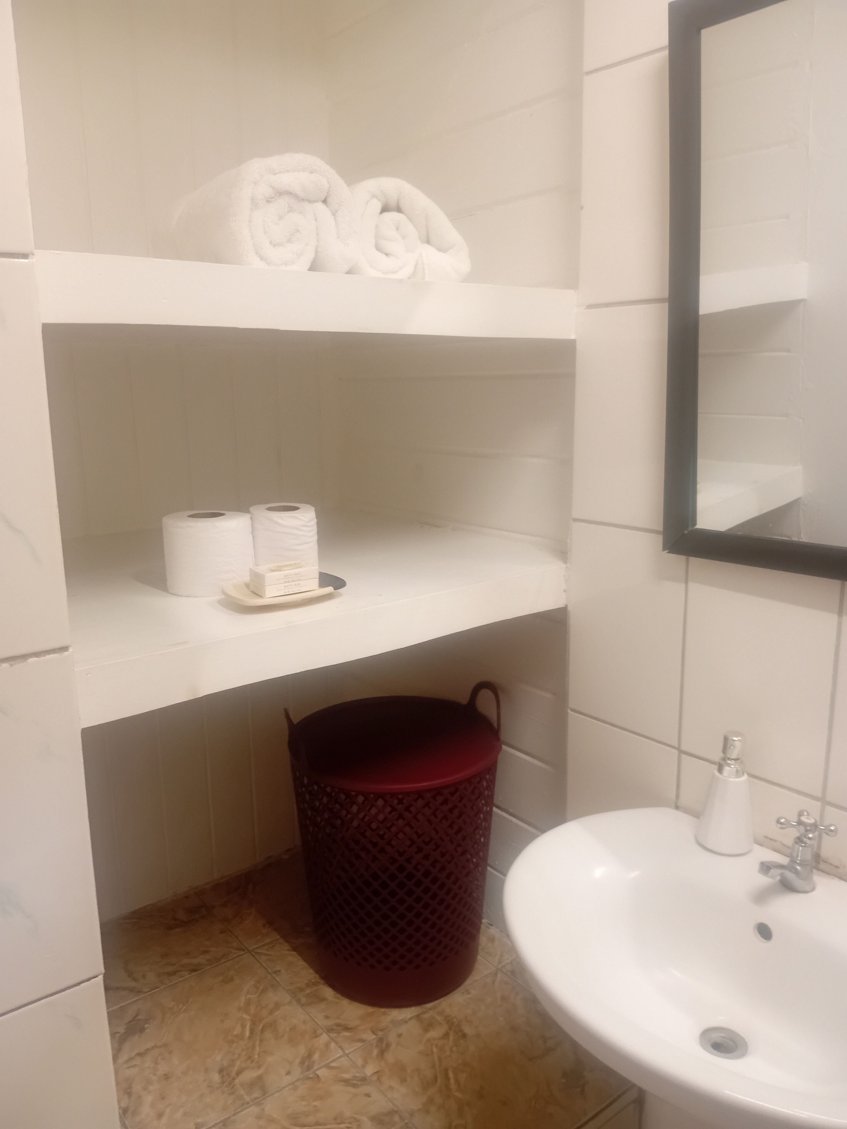 A bathroom with a white sink, a mirror, a red trash bin, and shelves holding towels and toilet paper.