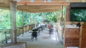 Un patio techado al aire libre con suelo de madera, mesas, sillas y una pantalla de televisión.