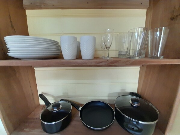 A kitchen shelf with stacked plates, white textured cups, wine glasses, and clear glasses. Below, there are three black pots with lids. - Pinard's Mountain Resort- Cottage 6 (Pointe Michel)