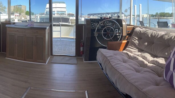 A boat interior with a wooden deck, a cushioned seating area, a steering wheel, and large windows offering views of the marina.