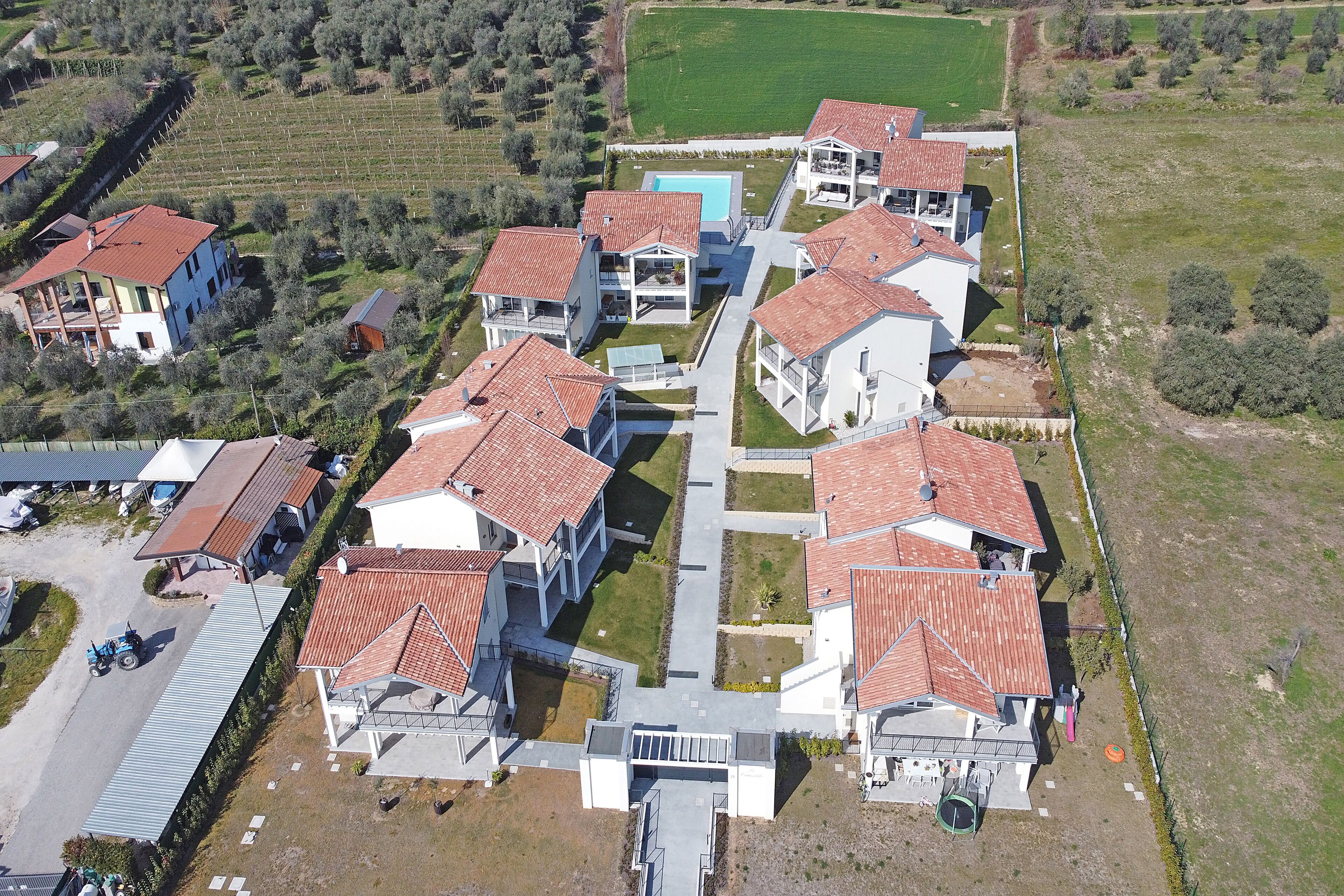 Vue aérienne d’un quartier résidentiel avec des maisons aux toits recouverts de tuiles rouges, une piscine centrale et des champs verdoyants environnants.