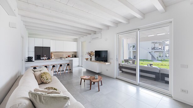 A modern living room with a sofa, a coffee table, and a TV. There is a kitchen area with cabinets and a dining table. - La Cittadella PT-6 by Wonderful Italy (Manerba del Garda)