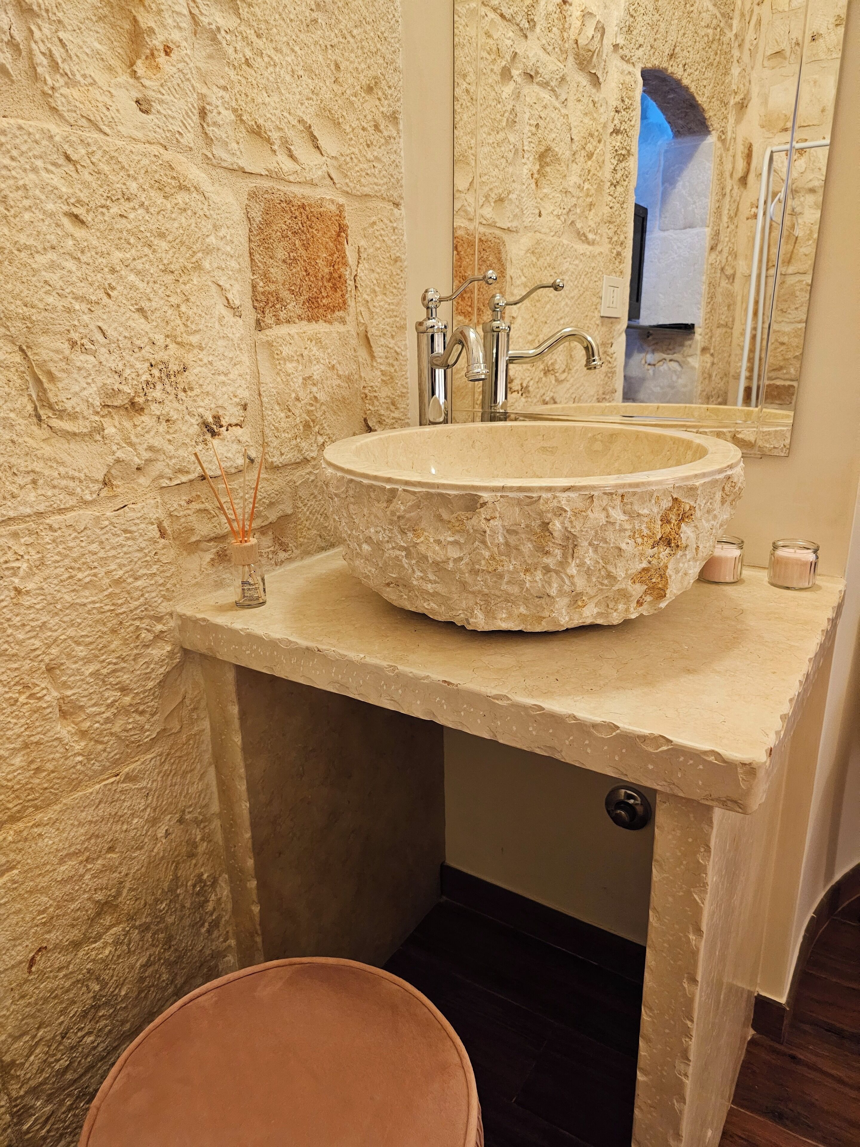 A stone sink with a mirror above it, a small candle holder, and a wooden stool.