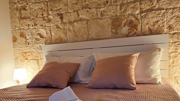 A neatly made bed with folded towels, a stone wall, and a white headboard. - PietraRosa Apartment (Triggianello)