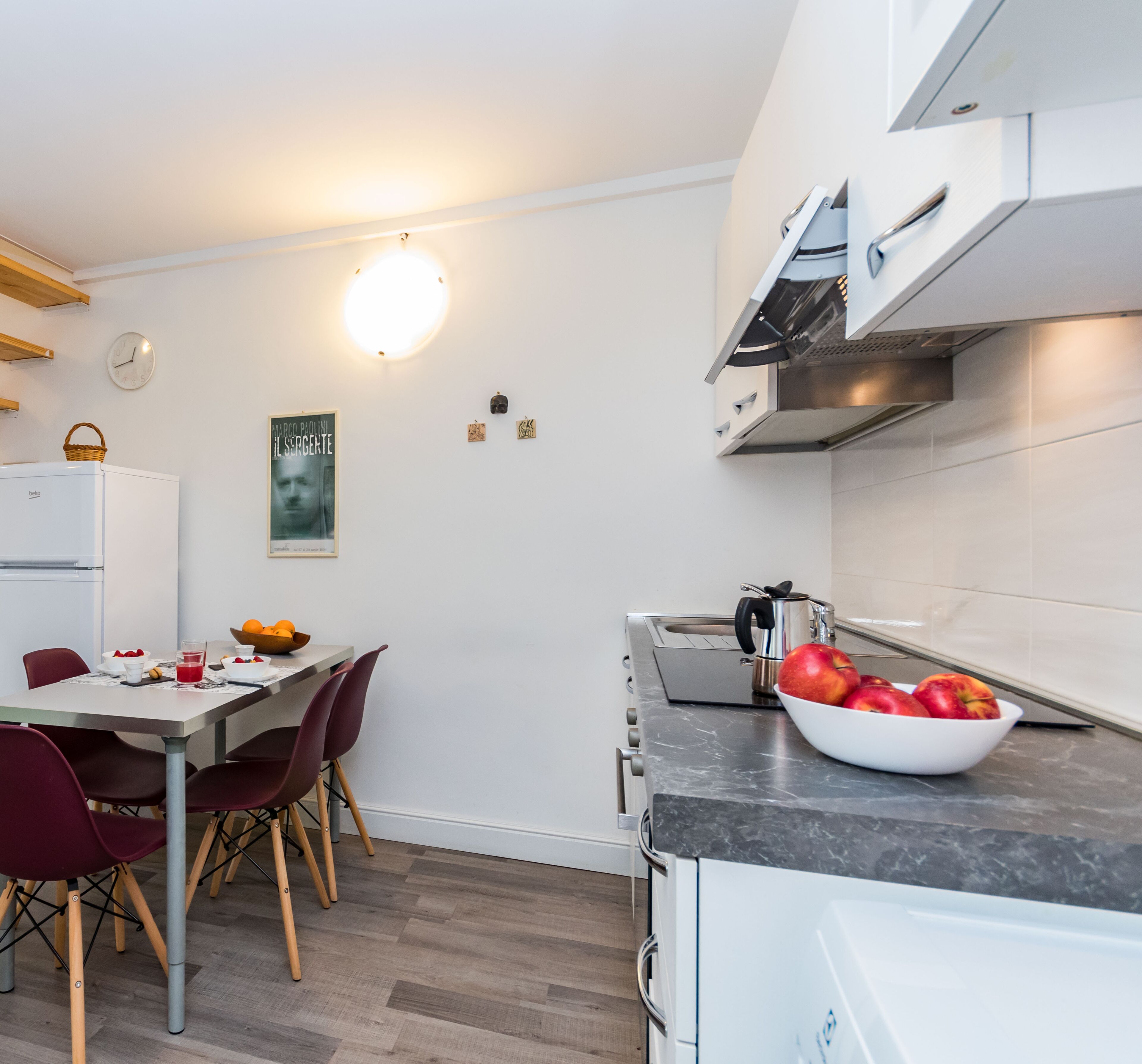 A modern kitchen with a dining area, featuring a white refrigerator, a sink, a stove, and a bowl of fruit on the counter.