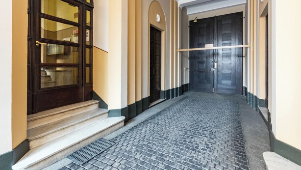 Un corridoio con pavimento in pietra, una scala che conduce a una porta in legno scuro e un'ampia finestra ad arco sul lato sinistro.