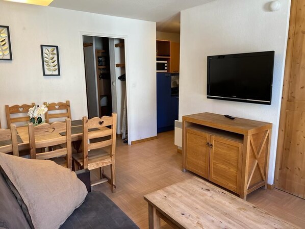 Un espace de vie compact comprenant une table à manger en bois, un canapé, un téléviseur fixé au mur et, au fond, une petite cuisine.