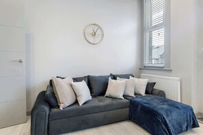 A modern living room with a grey sofa, a round wall clock, and a window with blinds.