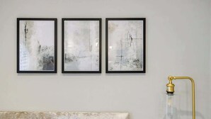 A wall with three framed abstract paintings and a clear glass lamp with a gold base. - Bright London Apartment with Central Heating (London)