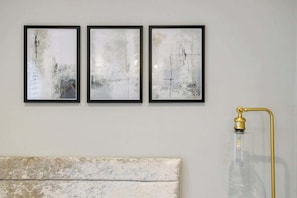 A wall with three framed abstract paintings and a clear glass lamp with a gold base.