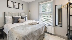 A bedroom with a bed, pillows, a window with blinds, a radiator, a bedside table, and a wall-mounted mirror. - Bright London Apartment with Central Heating (London)