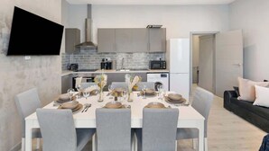A modern kitchen with grey cabinets, a flat-screen TV, and a dining area set for four with grey chairs and a white table. - Bright London Apartment with Central Heating (London)