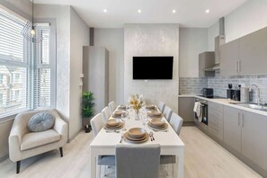 A modern kitchen with a dining area, a flat-screen TV mounted on the wall, and a window with blinds. - Bright London Apartment with Central Heating (London)