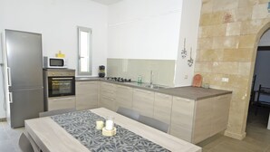 A modern kitchen with a built-in oven, microwave, and gas stove. There is a dining table with a patterned table runner and candles. The kitchen features light wood cabinets and a stone accent wall. - Villa Lara (Torre Pali)