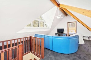 Una habitación con techo inclinado, un sofá azul, un televisor y una cama con manta blanca.
