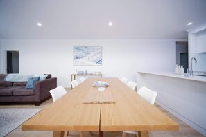 A modern dining area with a wooden table, white chairs, a painting on the wall, and a kitchen area with white cabinets. - Shoreacres Retreat - Your next holiday in paradise (Dunsborough)