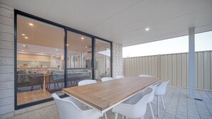 Une terrasse extérieure avec une table en bois et des chaises blanches, une porte-fenêtre coulissante donnant sur un espace intérieur et un sol carrelé.