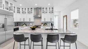 A modern kitchen with white cabinets, a central island with bar stools, stainless steel appliances, and a large window. - Gulf-front pool home in the heart of the Keys! Close to the beach! Guest house!  (Marathon)