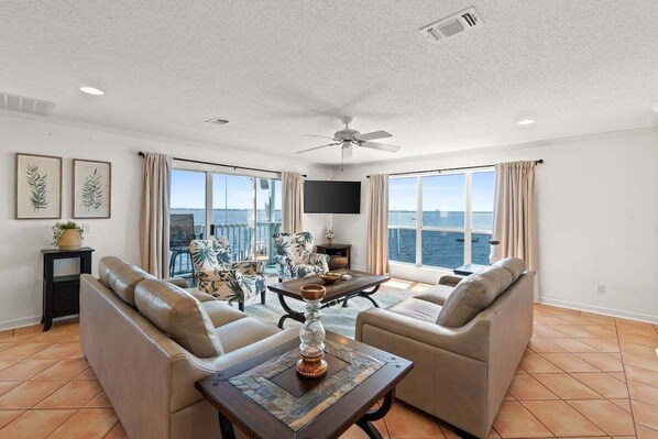 A spacious living room with a large window offering a view of the sea, a ceiling fan, and a flat-screen TV mounted on the wall.