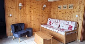 A room with a wooden wall, a sofa with patterned cushions, a dark armchair, and a small wooden table.