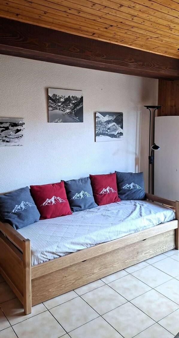 A wooden sofa with a quilted cover and mountain-themed pillows, a wall with framed photographs, and a floor lamp.