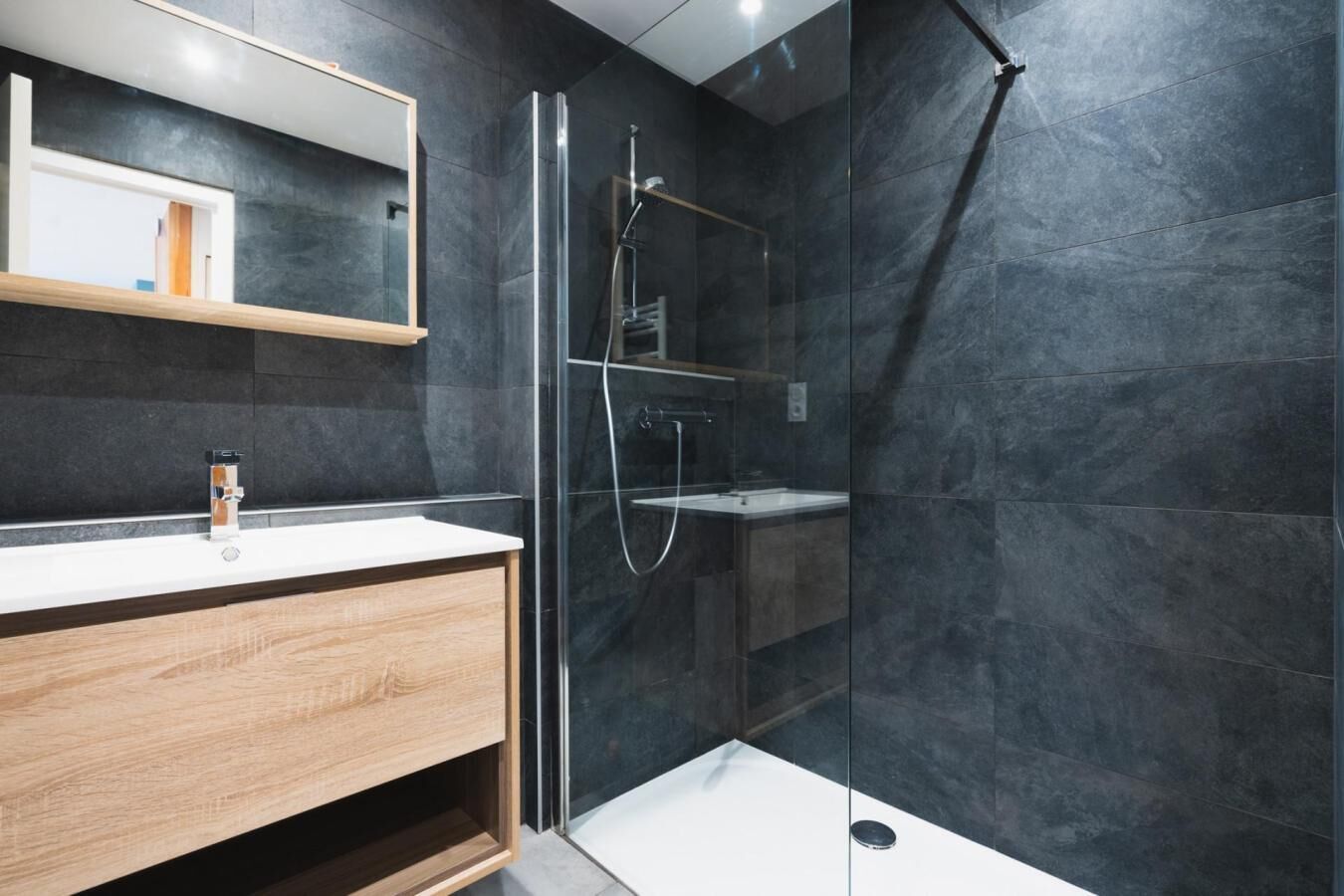 Une salle de bain moderne avec une douche à parois de verre, un meuble-lavabo en bois et des murs recouverts de carreaux foncés.