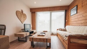 Uma sala de estar aconchegante com um sofá de madeira, uma mesa de centro, uma televisão sobre um suporte de madeira e uma janela grande com cortinas.