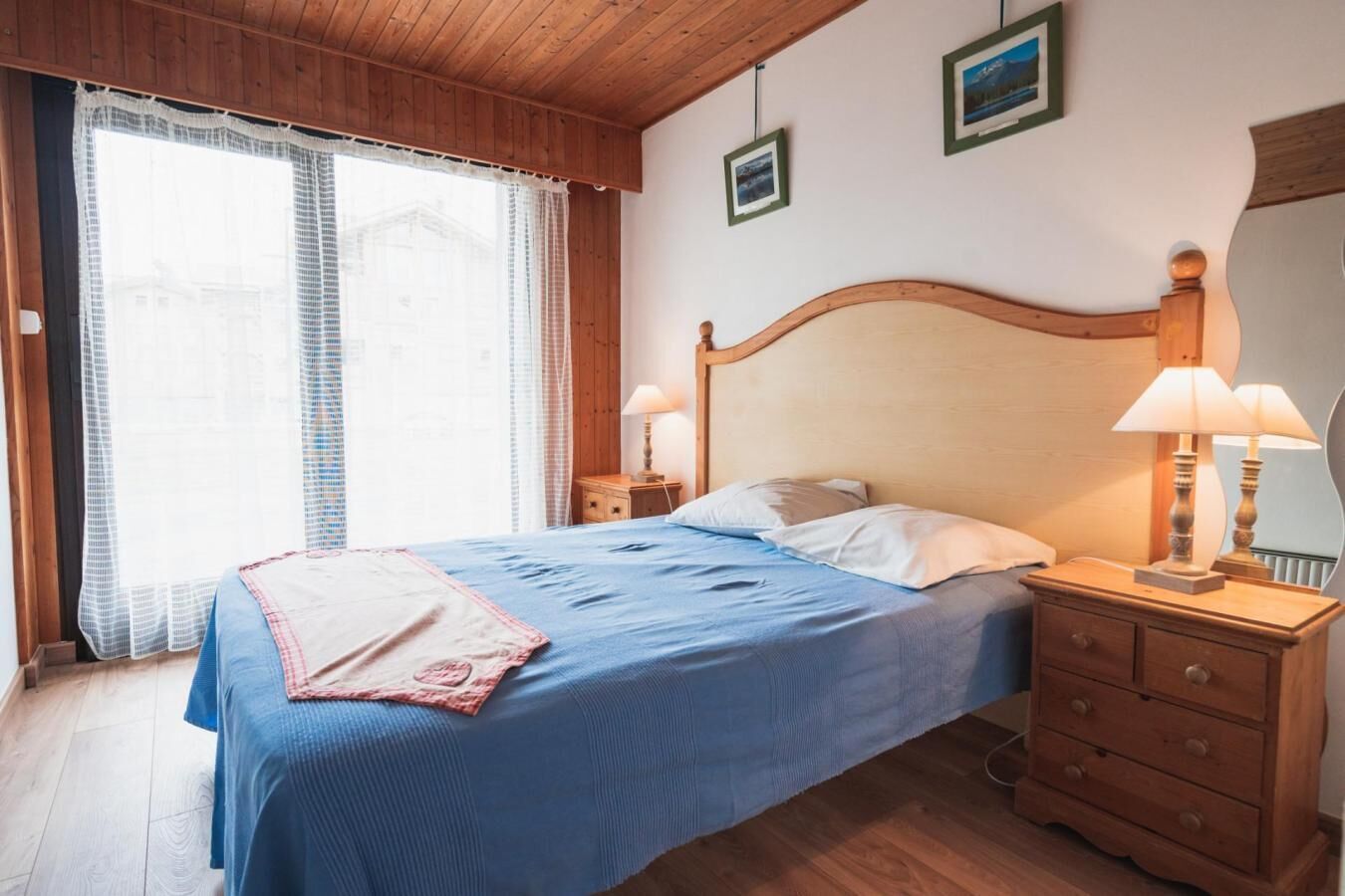 Um quarto com cama de madeira, duas mesas de cabeceira com luminárias, uma janela com cortinas e um pequeno aparador de madeira.