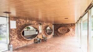Um hall amplo com piso de tijolos e arcos circulares. Há balcões de recepção e uma área de estar com uma planta em vaso.
