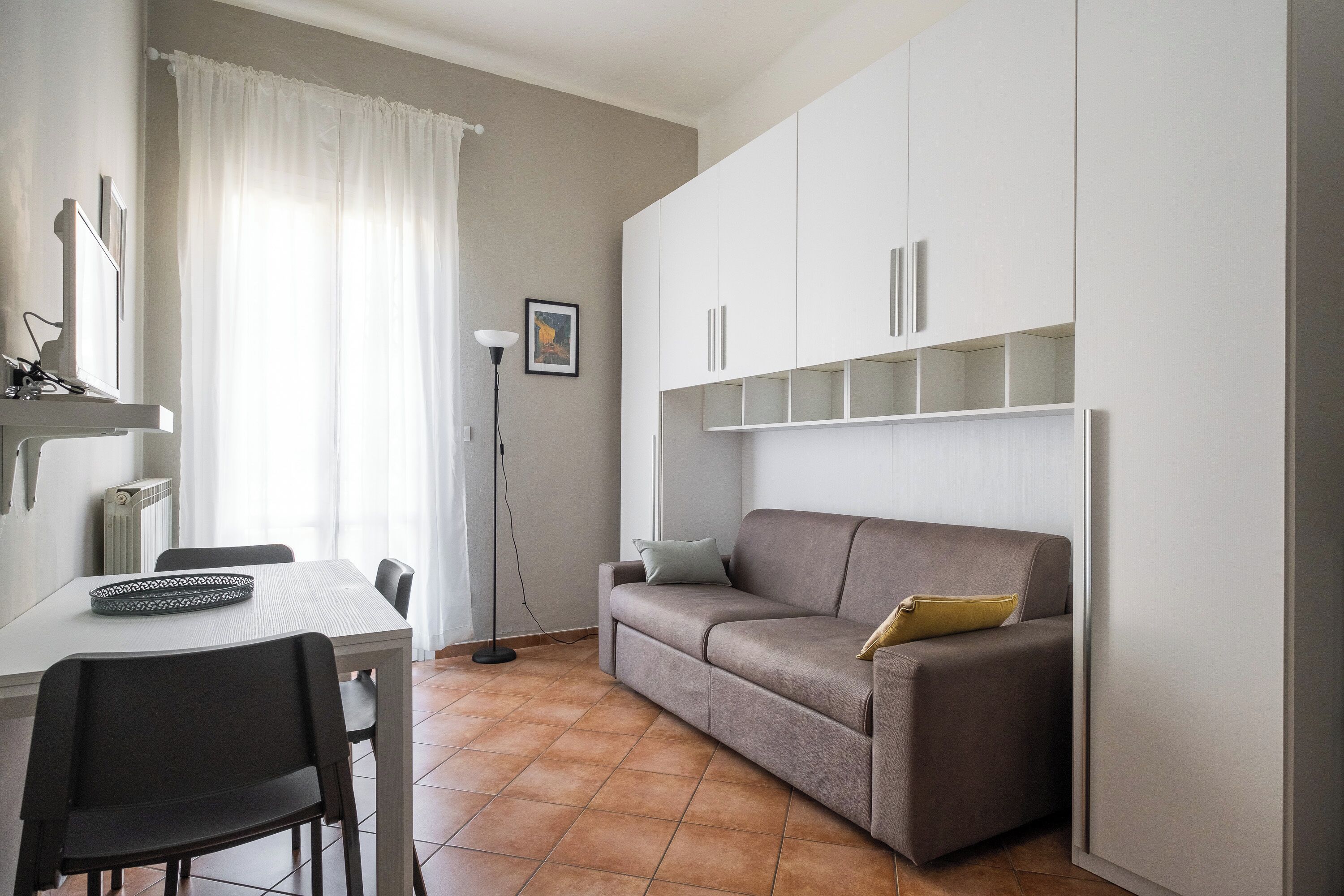 A modern living room with a sofa, a dining table, and chairs. There is a radiator, a floor lamp, and a framed picture on the wall.