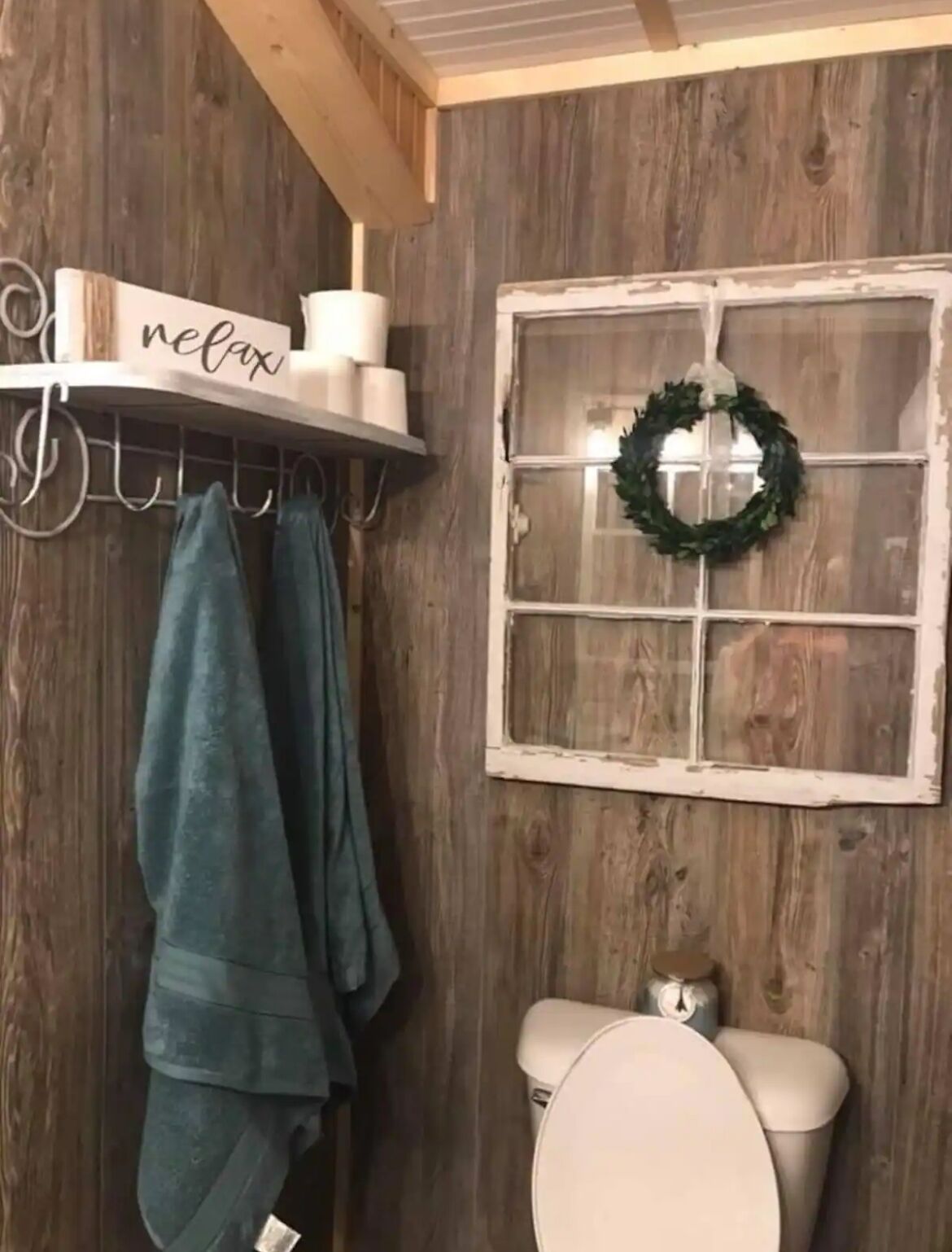 Een rustieke badkamer met houten wanden, een plank met toiletartikelen, een raam met een krans en handdoeken aan haakjes.