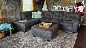 A cozy living room with a sectional sofa, a small table, and a decorative wall with a compass and a sign that reads "The Cabin". - Charming waterfront barndominium on the beautiful spring-fed Chipola River. (Altha)