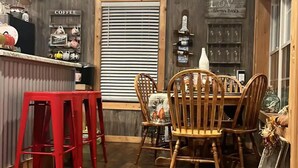 A cozy dining area with a wooden table and chairs, a red stool, and a coffee station with various mugs and a sign that reads "COFFEE". - Charming waterfront barndominium on the beautiful spring-fed Chipola River. (Altha)