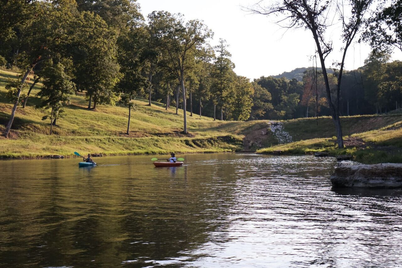 A calm river with kayaks, surrounded by trees and grassy banks.