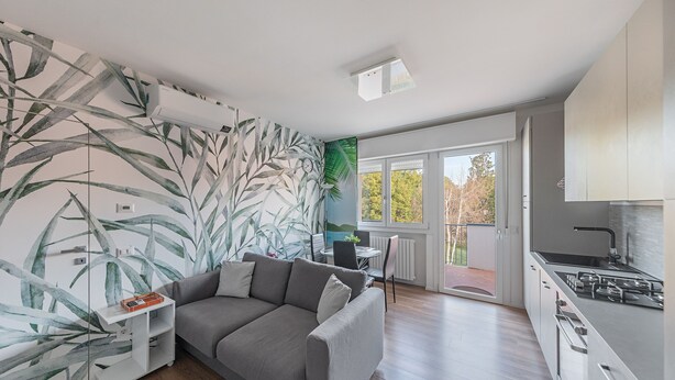 A modern living room with a grey sofa, a small white side table, and a wall with a tropical leaf mural. - Maisonnette Il Todeschino B12 by Wonderful Italy (Sirmione)