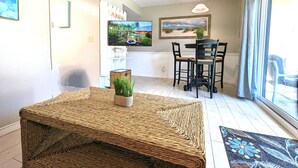 Un salon avec une table en osier, un téléviseur fixé au mur et un coin repas comprenant des chaises et une table.