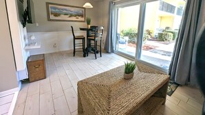 Une pièce comprenant une table à manger, des chaises, un ventilateur de plafond, une grande fenêtre avec des rideaux et un banc en osier agrémenté d’une plante.