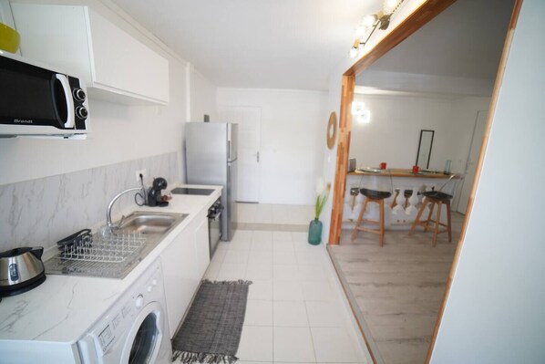 A compact kitchen with a microwave, sink, and a washing machine. There is a dining area with a table and chairs visible through an open doorway. - Edith T2 design, la plage à 200m
(Sainte-Luce)