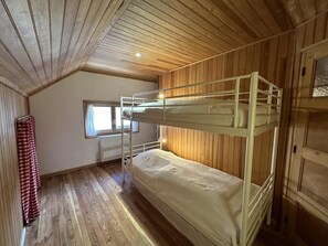 A bunk bed room with wooden walls and floors, a window with curtains, and a radiator.